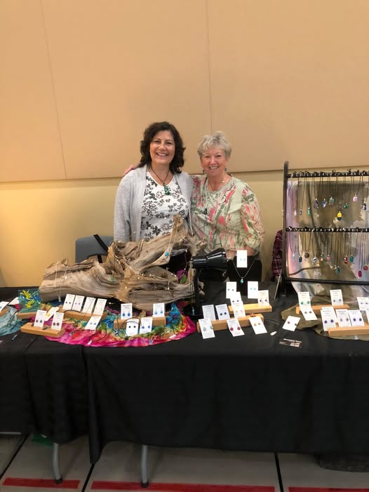 Two women smiling behind a table of handmade glass jewelry.